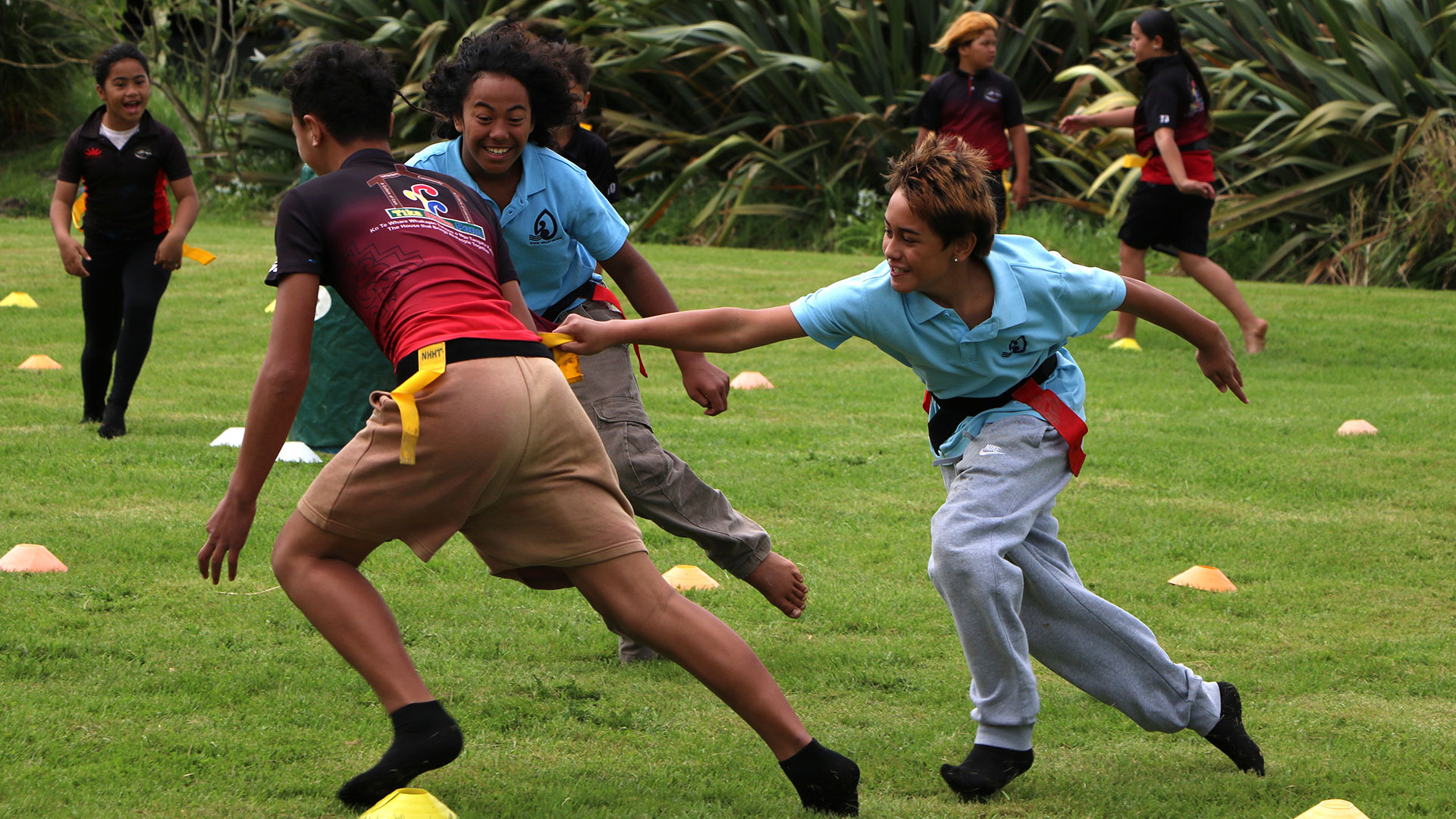 Children play a fast‑paced game of Ki‑o‑Rahi on a grassy area at Te Houhanga a Rongo Marae, running between cones while passing and defending a tag belt as others move into position nearby.