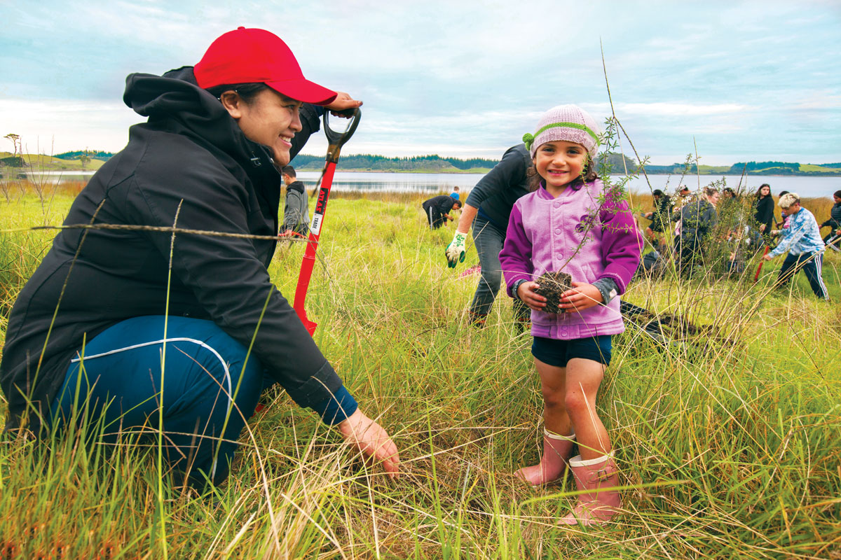 Exterior, near harbour -woman holding a spade crouches next to a young girl at a planting day.
