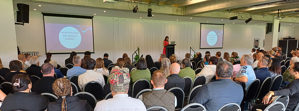 Many people seated in a room watching and listening to a woman presenting on a stage.