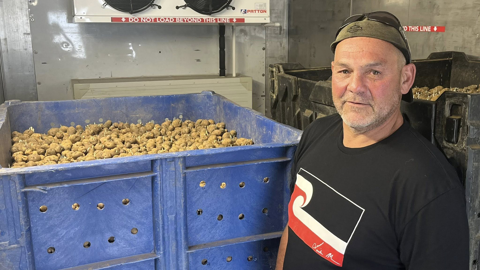 Man wearing a cap backwards and black t-shirt standing in front of a large blue bin of potatoes.