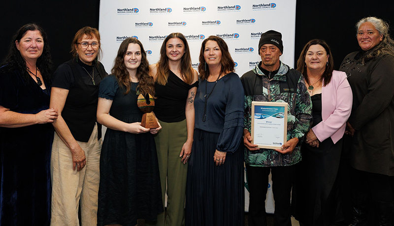 Seven women and one man (wearing a beanie) with a trophy and certificate in front of Northland Regional Council logo backdrop. (Photo: Dawn Dutton Photography).