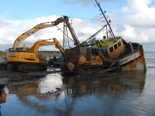 Diggers on beach recovering wrecked boat.