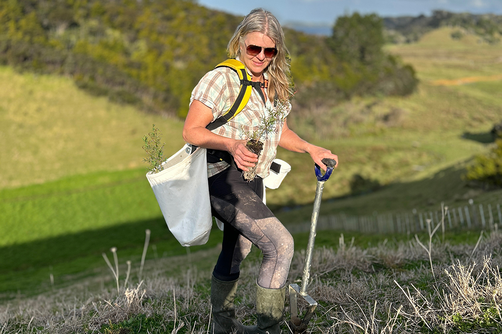 Woman with seedlings and planting equipment on hilly farmland.