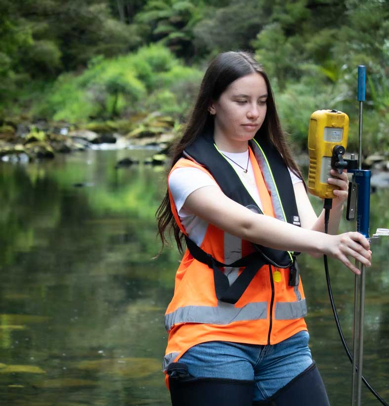 Woman standing in river wearing orange vest and life jacket holding hydrology recording equipment.