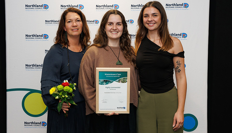 Three women with bouquet of flowers and certificate in front of Northland Regional Council logo backdrop. (Photo: Dawn Dutton Photography).