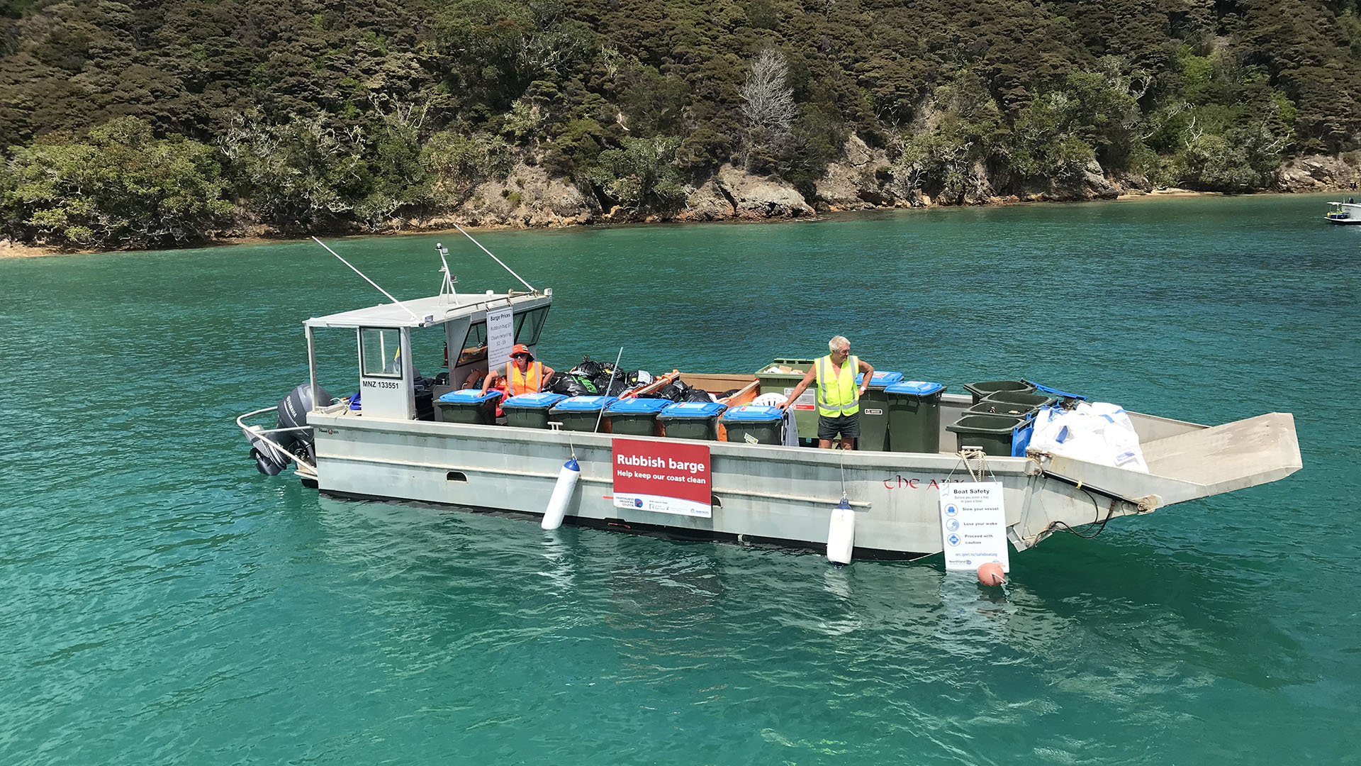 A grey barge with two people and blue-top bins on the sea water.