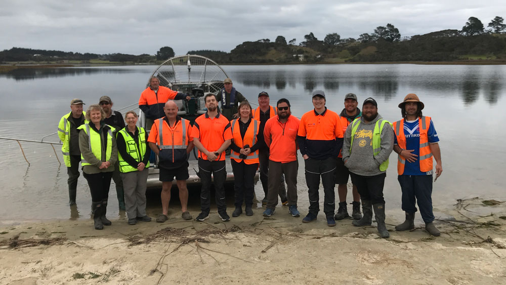 A group of people wearing hi-vis clothing standing by Lake Ngatu, in front of a lake boat.