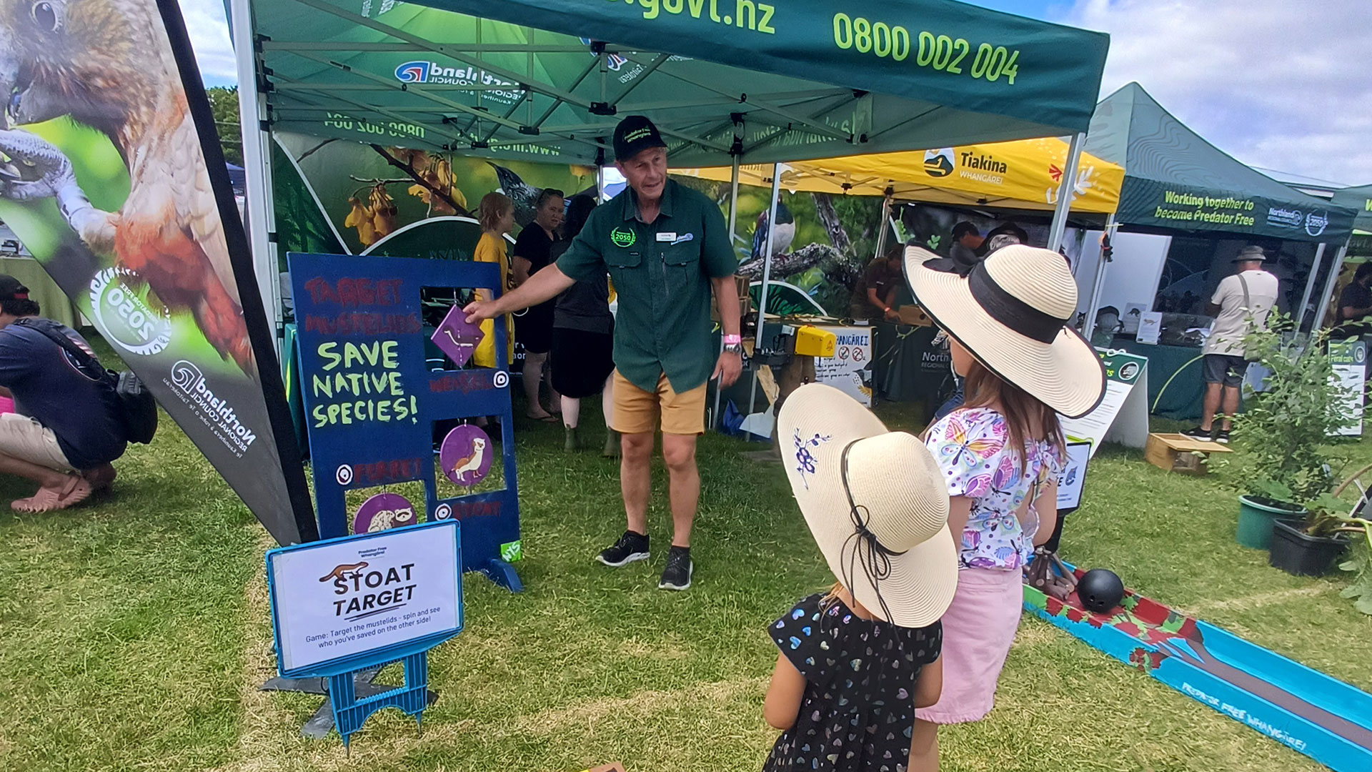 Two girls wearing sun hats talking to a man standing near an interactive game.