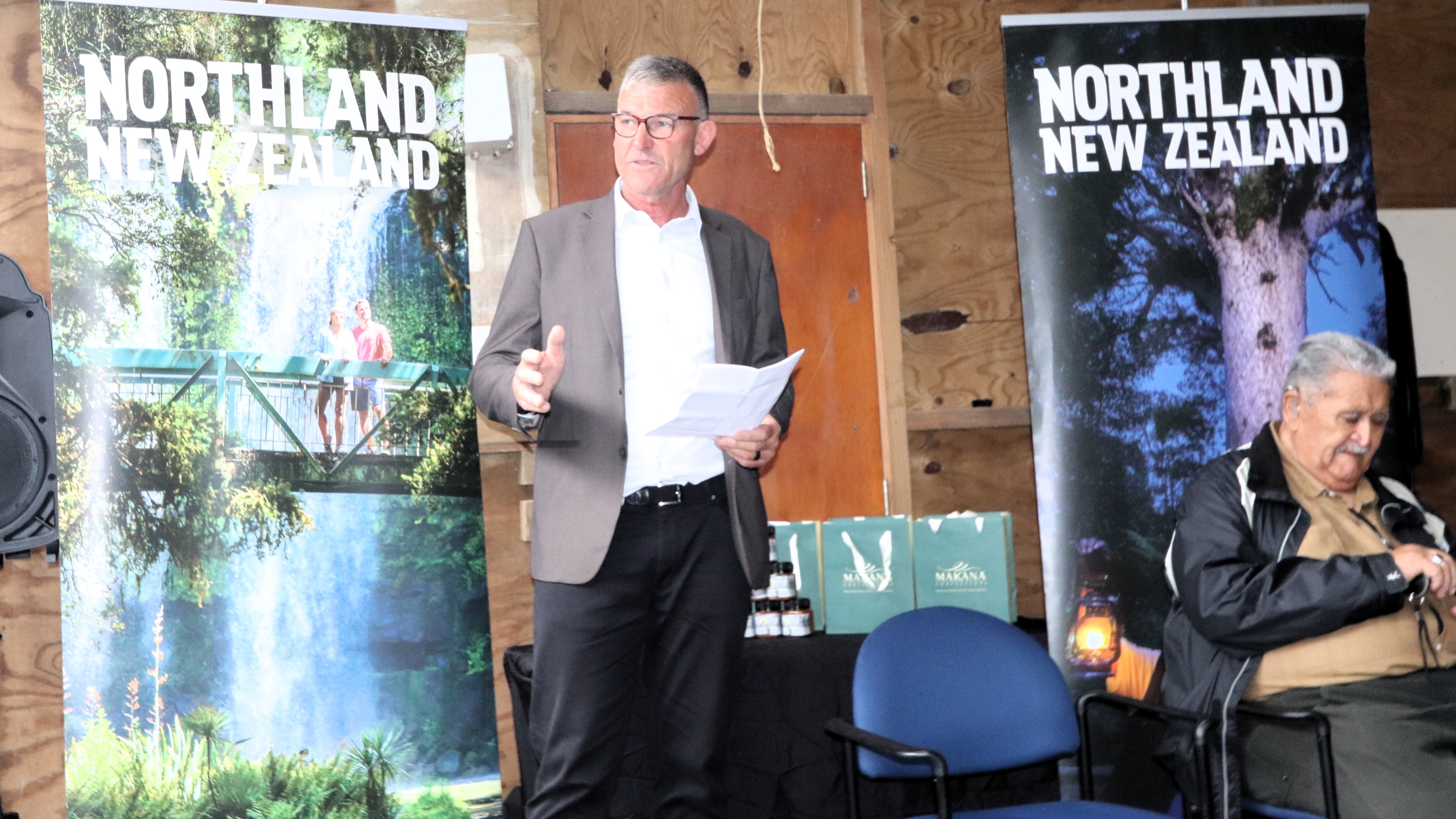 Northland Regional Council member Geoff Crawford stands indoors holding papers and speaking, positioned between two tall banners reading “Northland New Zealand,” one showing a forest waterfall and bridge and the other a large tree. Nearby are chairs and a small table with gift bags, set against wooden walls in a simple event space.