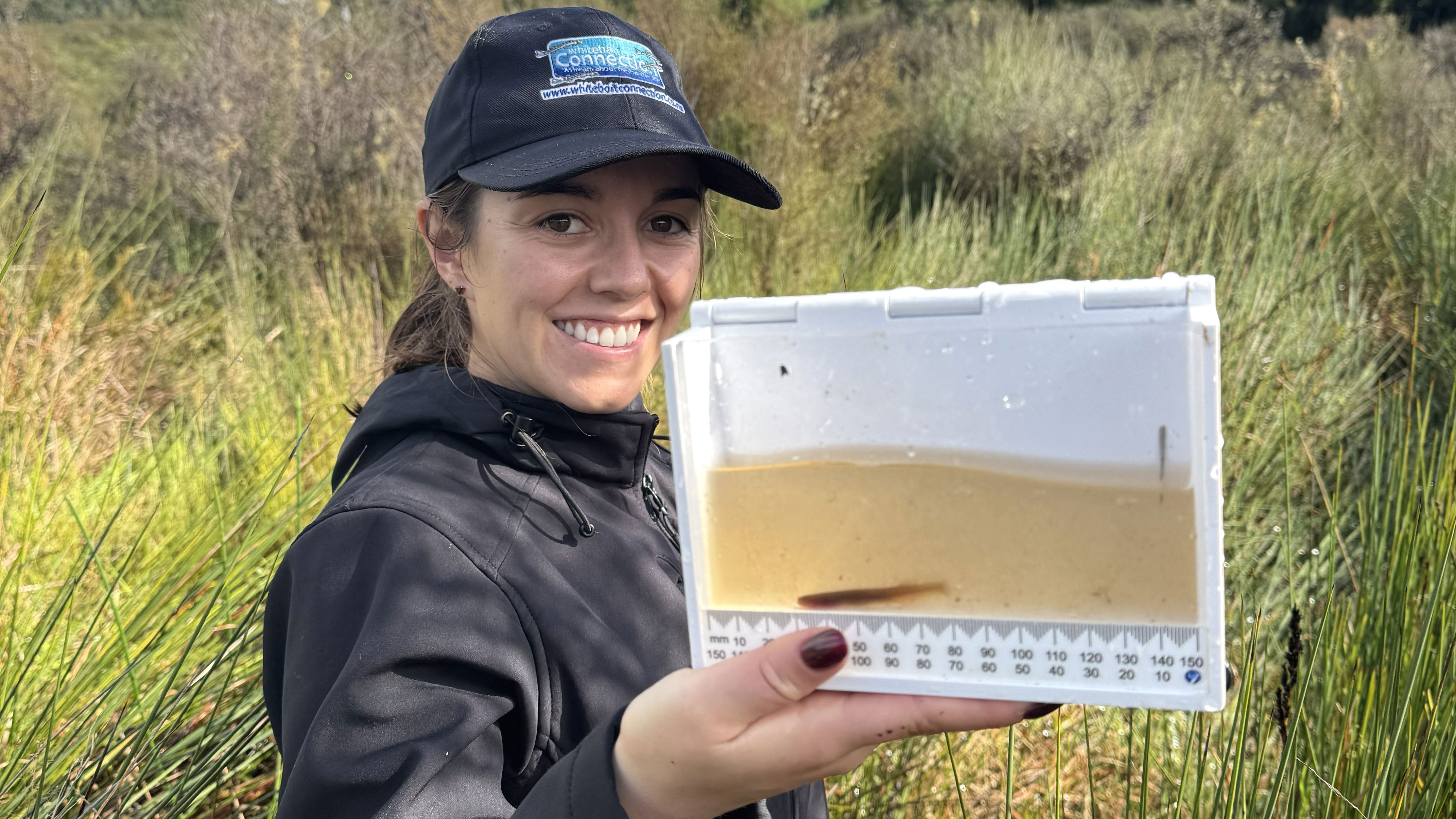 A woman wearing a cap, smiling and holding a small measuring tank with a mudfish in water.
