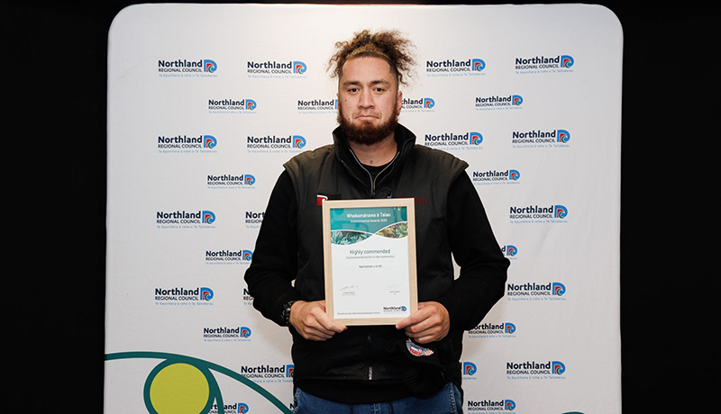 Man wearing black clothing holding a certificate in front of Northland Regional Council logo backdrop. (Photo: Dawn Dutton Photography).