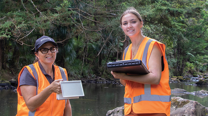 Two woman wearing orange vests holding a clipboard and device with river, rocks and bush in background.