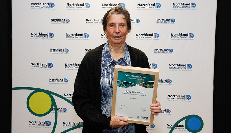 Woman wearing black cardigan holding her certificate in front of Northland Regional Council logo backdrop. (Photo: Dawn Dutton Photography).