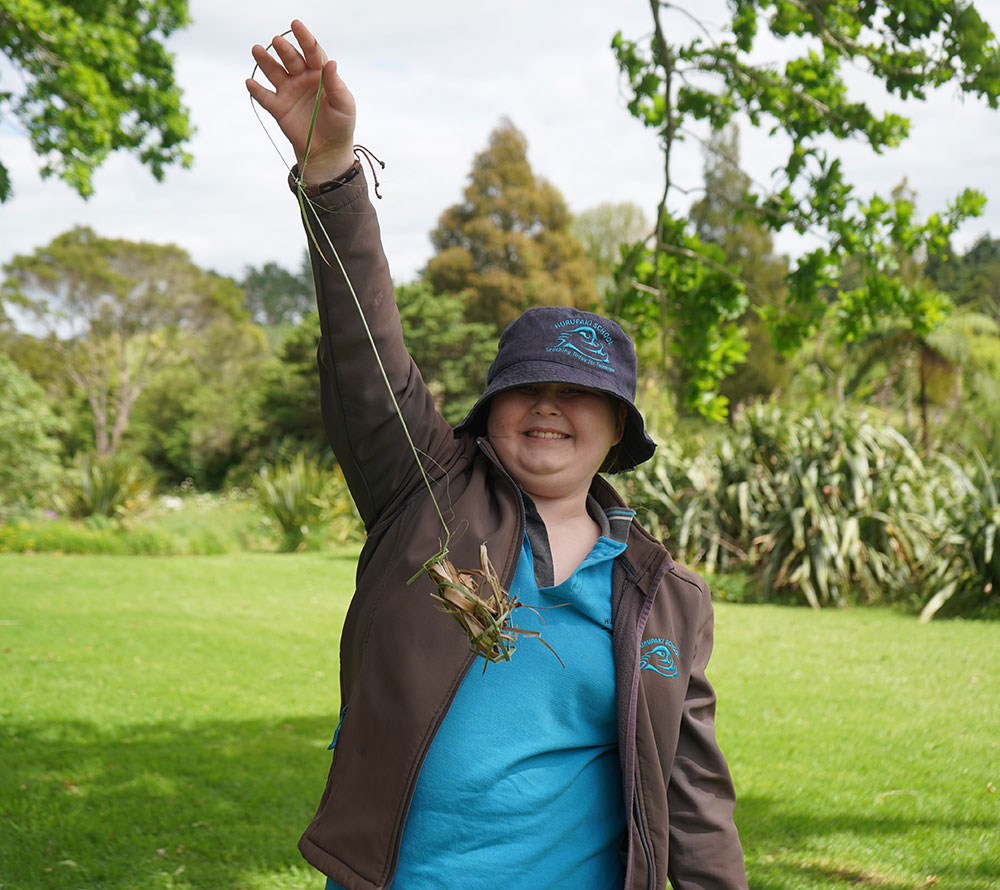 : A young female stands outdoors at Pehiāweri Marae, holding up a handmade poi toa crafted from tī kōuka leaves, with grass and trees visible in the background.