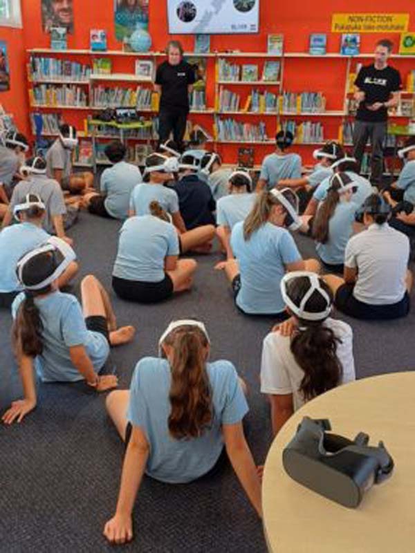 group of students wearing virtual reality headsets sit on the carpeted floor of a library, two adults stand near bookshelves.