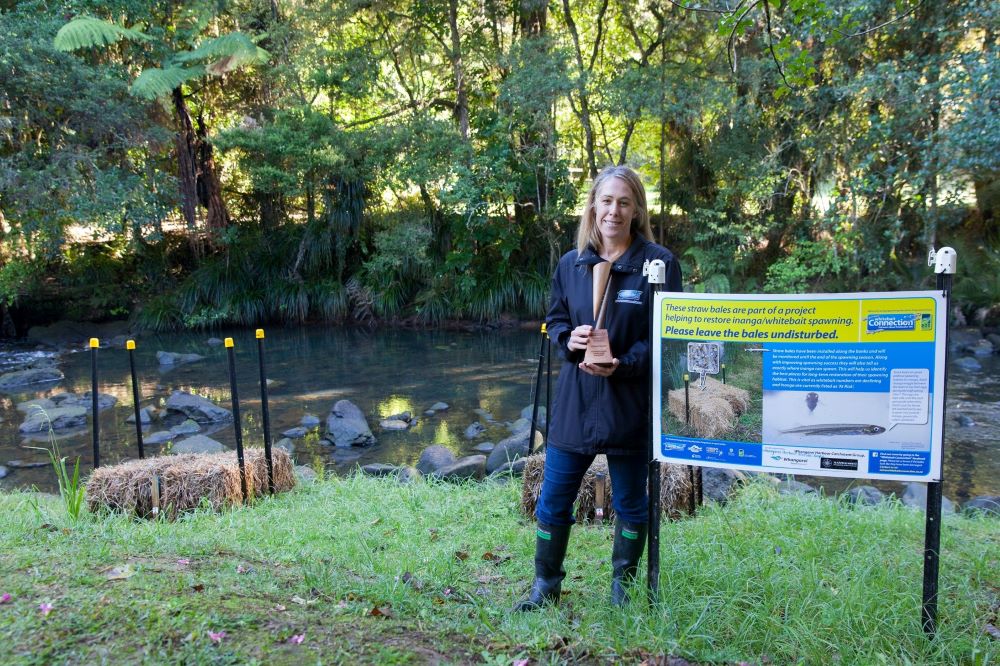 Woman holding a trophy by sign next to a stream.