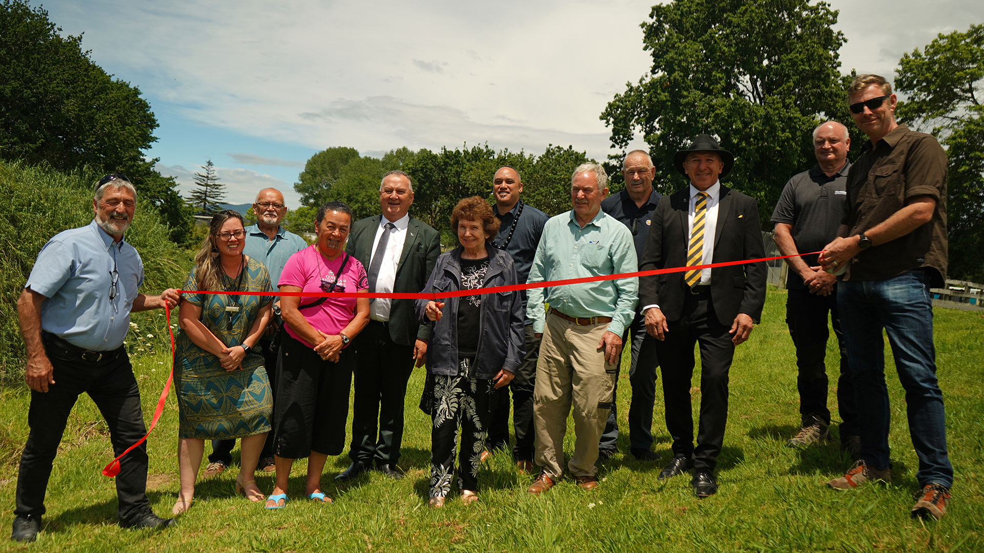 A group of people in a line with a red ribbon across the front of the group. A woman in the centre is about to cut the ribbon with scissors.