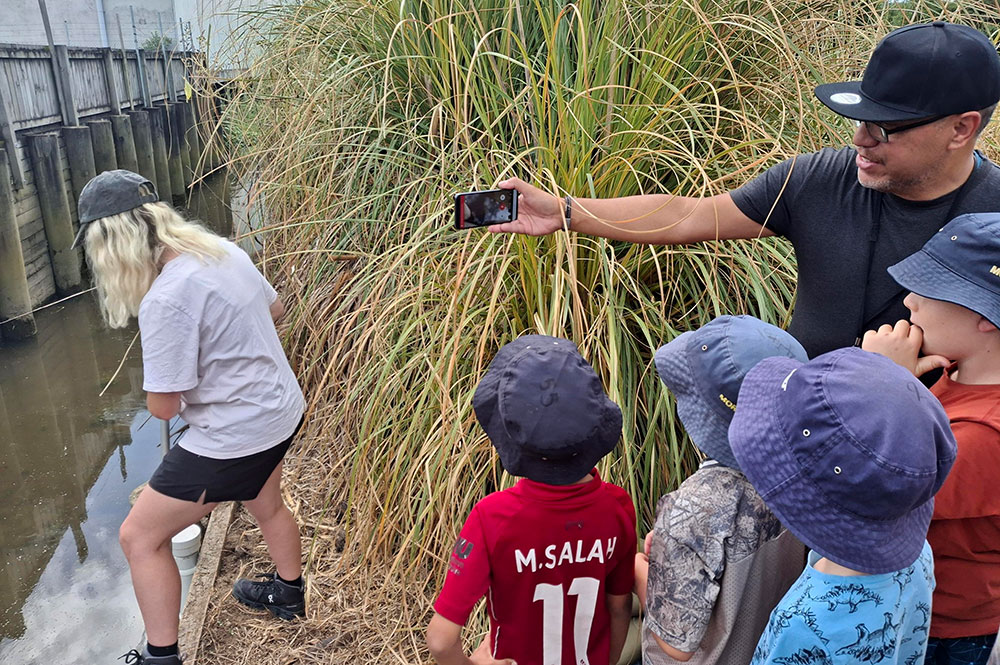 Four children looking at a phone being held by an adult male. Adult female put camera in nearby water.
