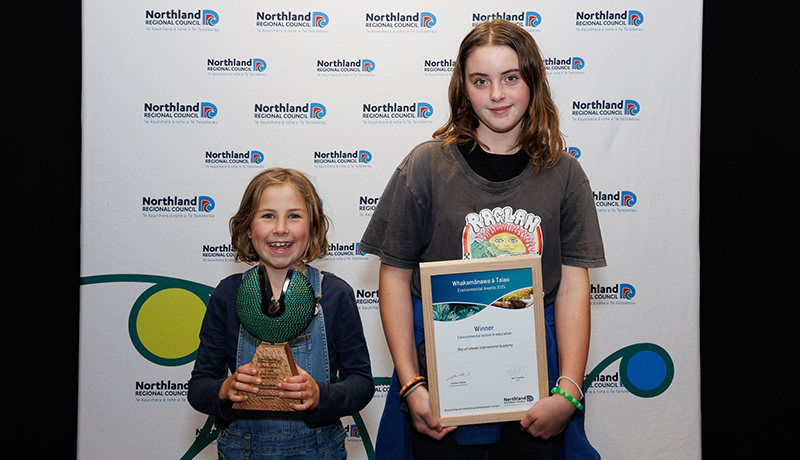 Two female students with their trophy and certificate in front of Northland Regional Council logo backdrop. (Photo: Dawn Dutton Photography).