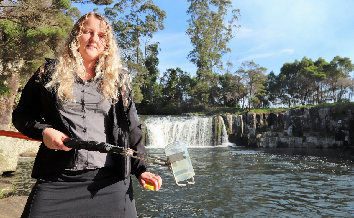 Woman holding water sampling pole in front of waterfall.