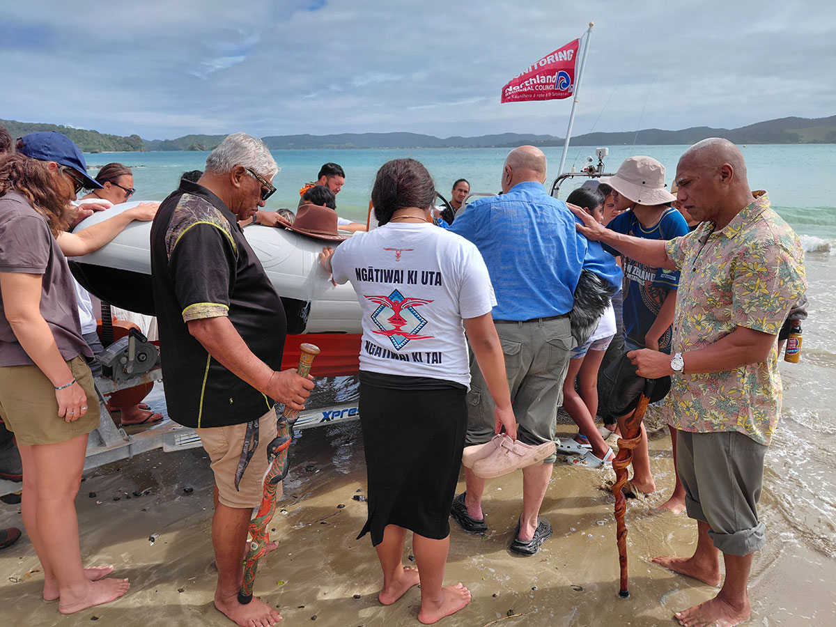 People with their hands placed on the inflatable boat as it is named and launched at a beach.