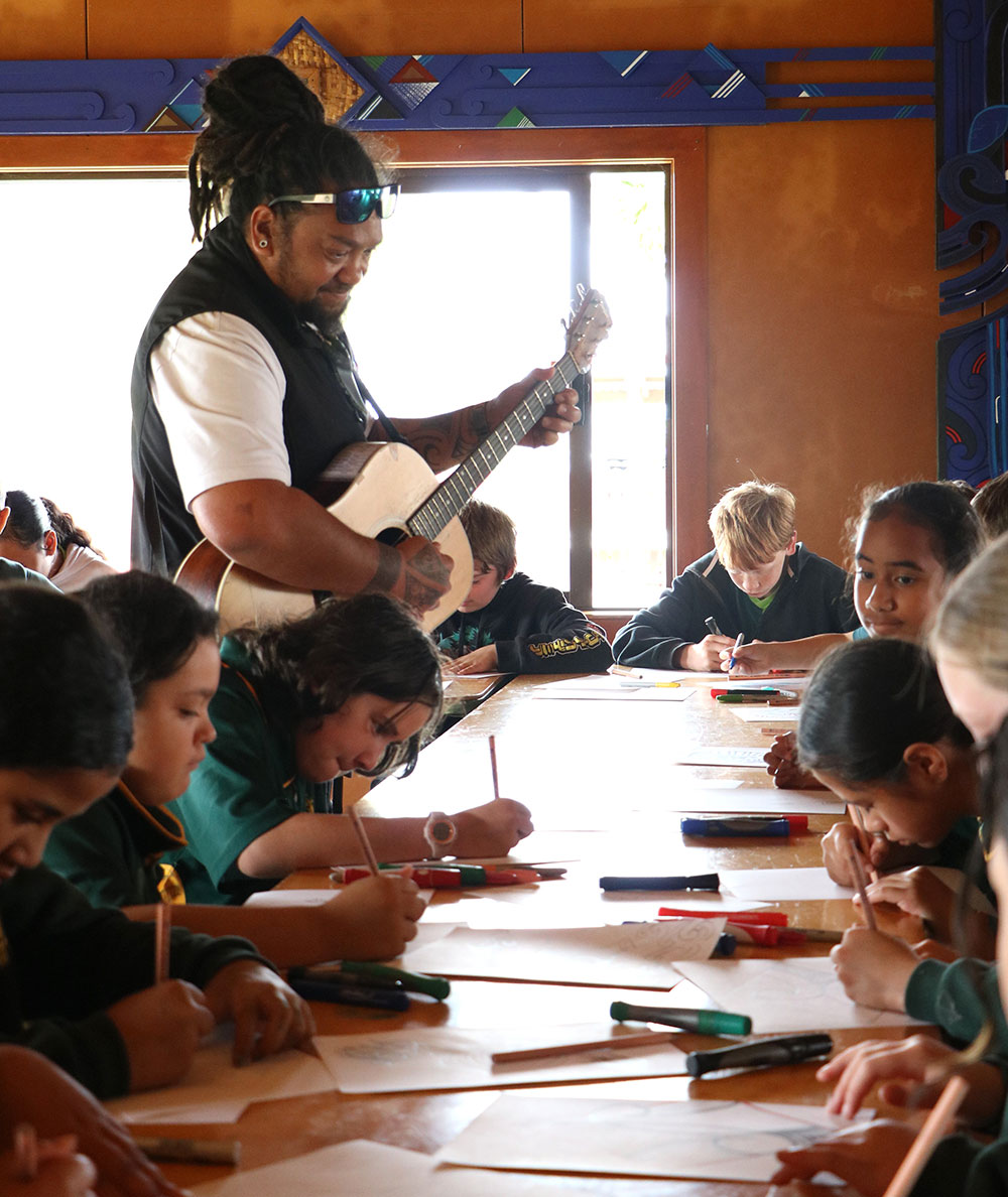 Children sit around a table inside, drawing carving patterns with pens and paper while a facilitator stands nearby playing a guitar and supporting the learning.