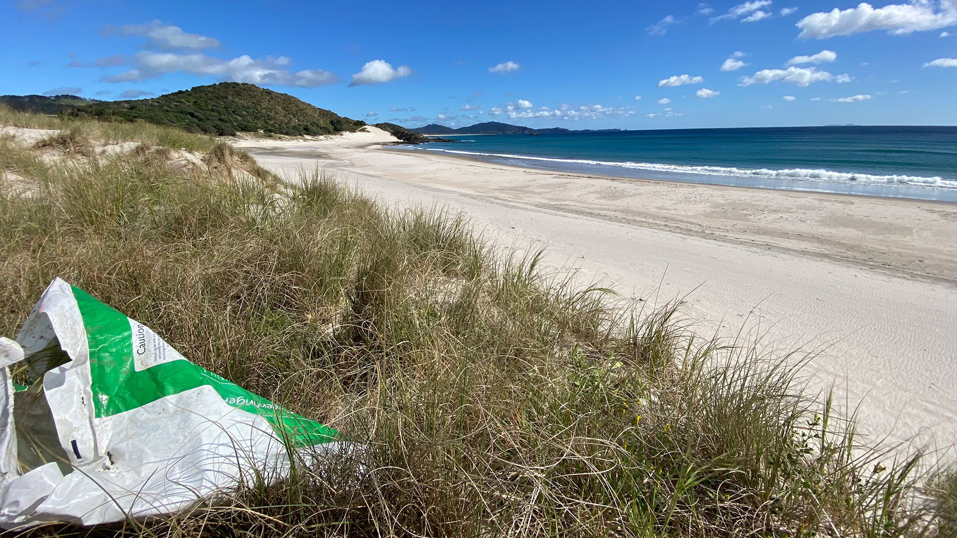 View down the beach of sand dunes, ocean, sky and hills in the background, with a weed sack in the foreground.