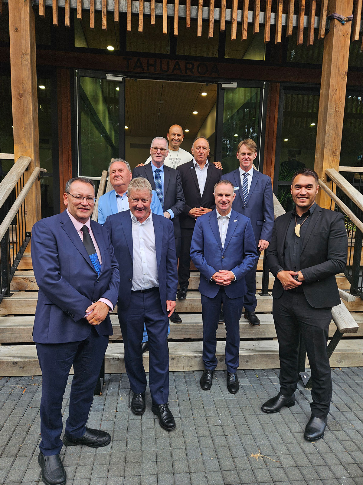 A group of 9 men wearing suits stand in front of a building.