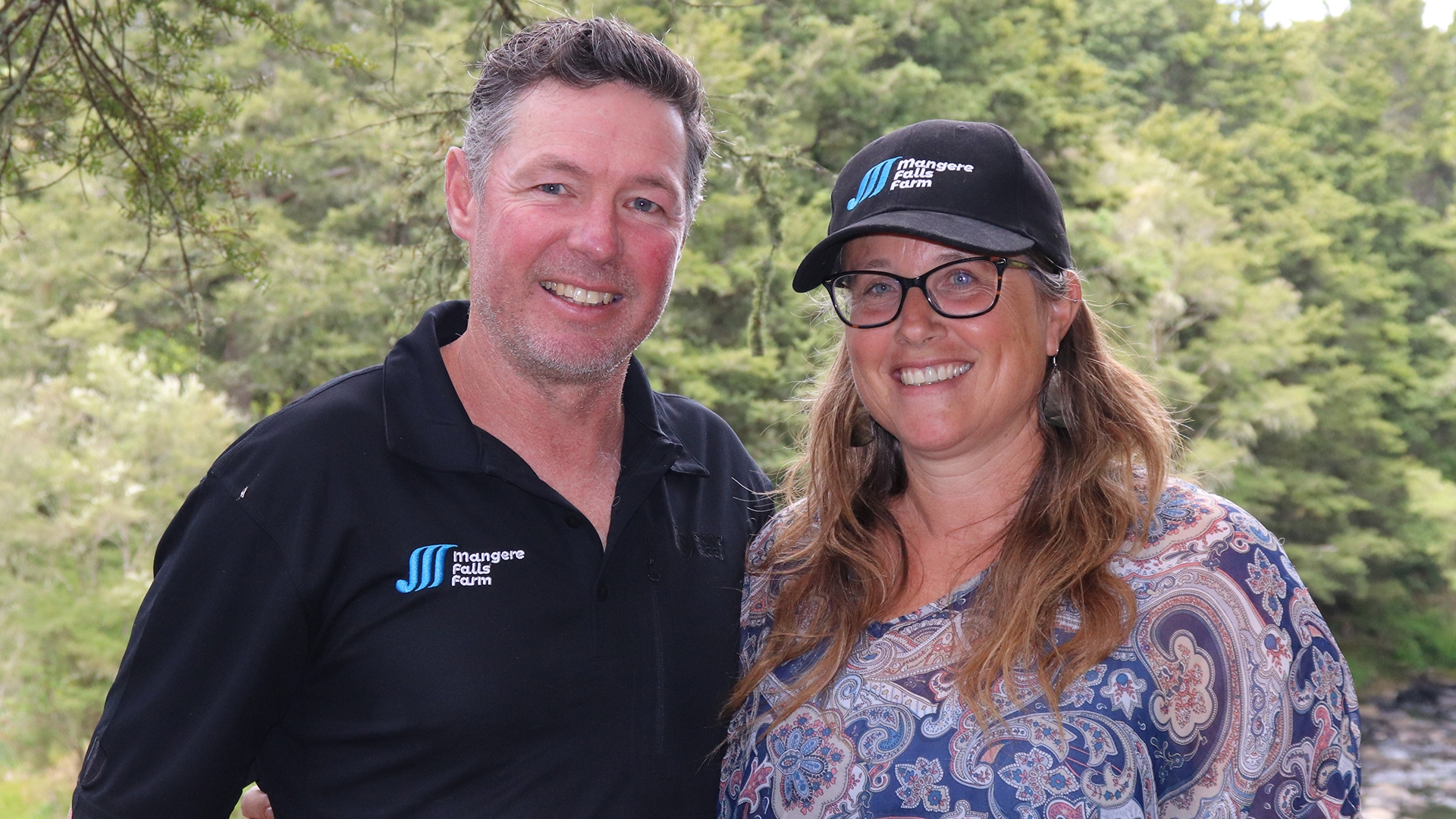 Man and a woman wearing a cap standing in front of trees.