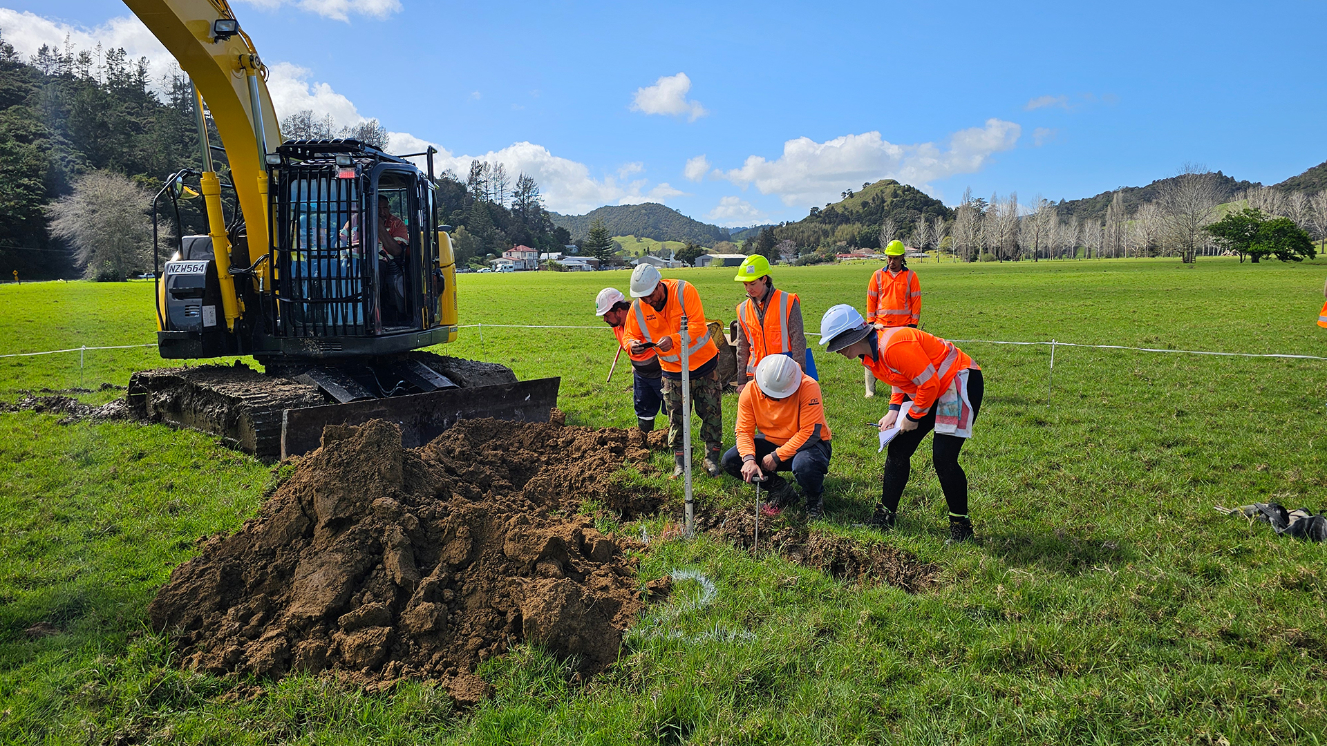 People wearing hi-vis vests and hard hats look into a trench beside a digger.
