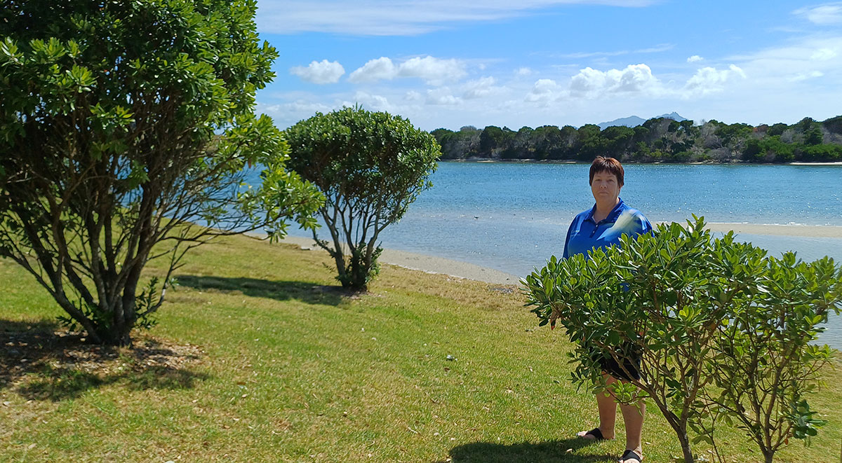 A woman stands behind small native tree. with estuary in the background.